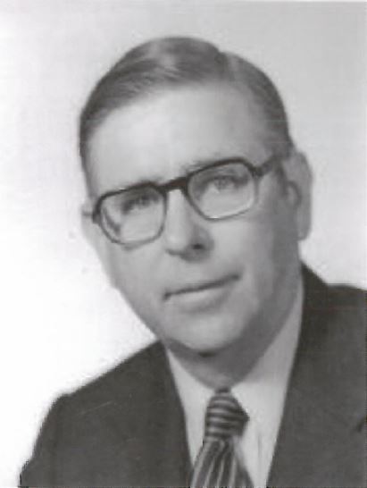 Black and white vintage headshot portrait of a man wearing dark-rimmed eyeglasses, a suit jacket, dress shirt, and striped tie.