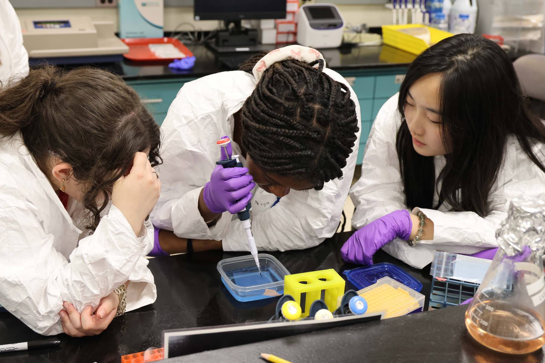 Three students in lab coats work together, with one using a micropipette to dispense fluid into a container on a laboratory bench.