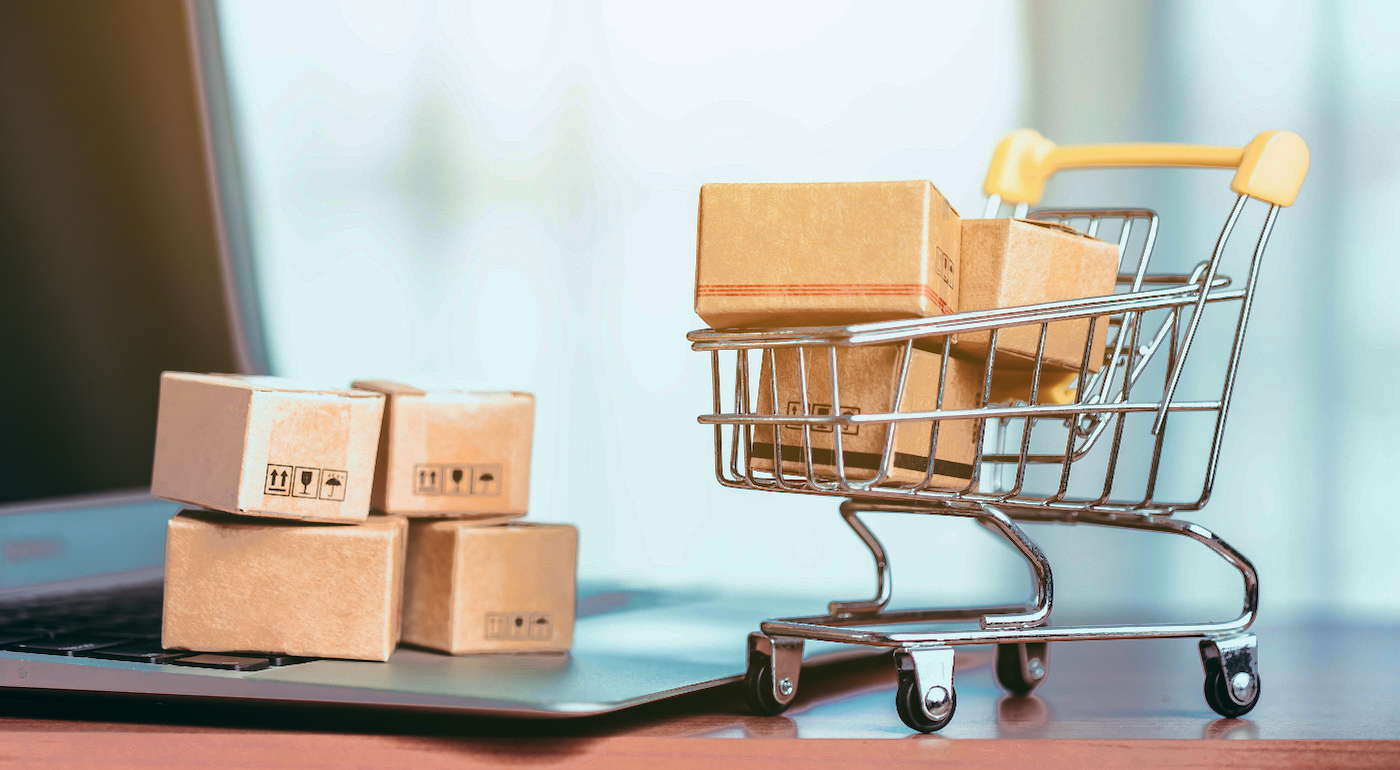 Small cardboard boxes stacked on a laptop keyboard next to a miniature toy shopping cart filled with more packages.