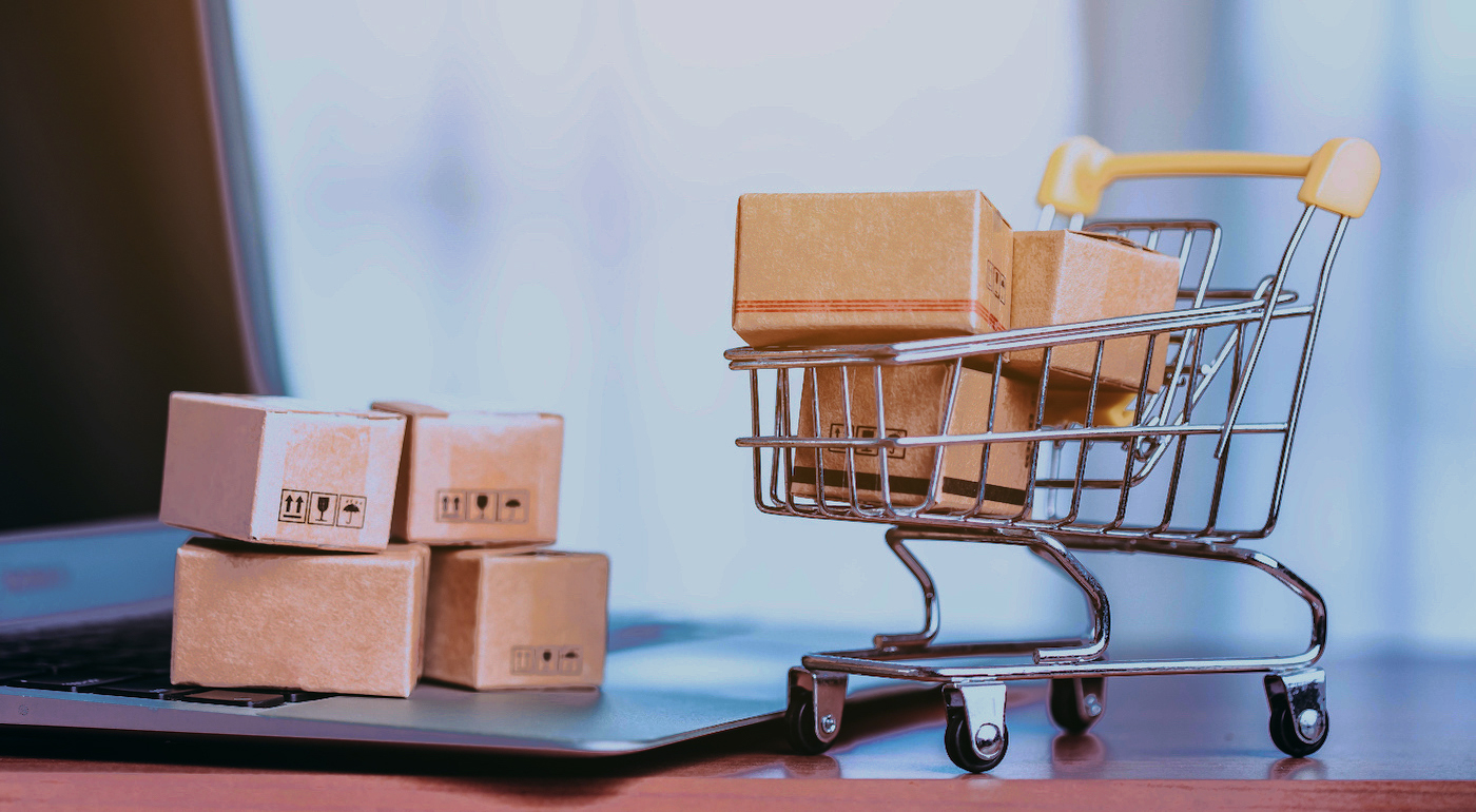 Small cardboard boxes stacked on a laptop keyboard next to a miniature toy shopping cart filled with more packages.