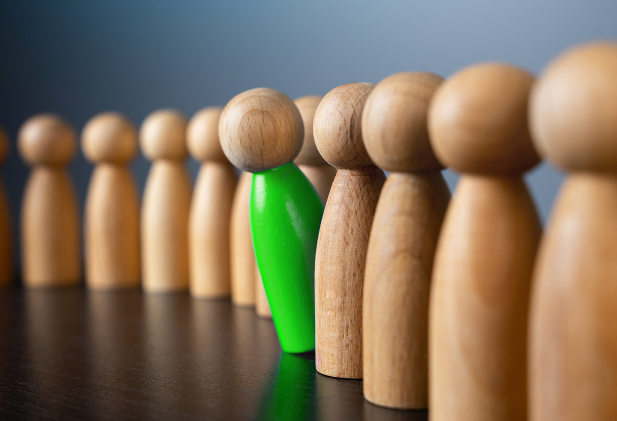 A row of wooden peg figures on a dark surface, with one figure in the middle painted bright green to stand out from the rest