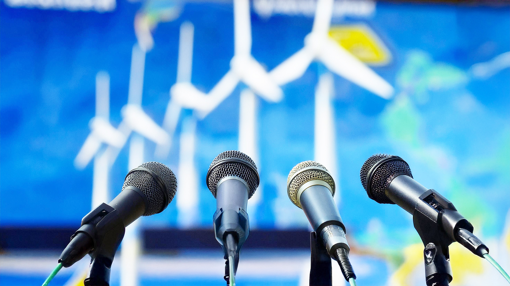 Four microphones lined up on stands for a press conference with a blurred backdrop of wind turbines.