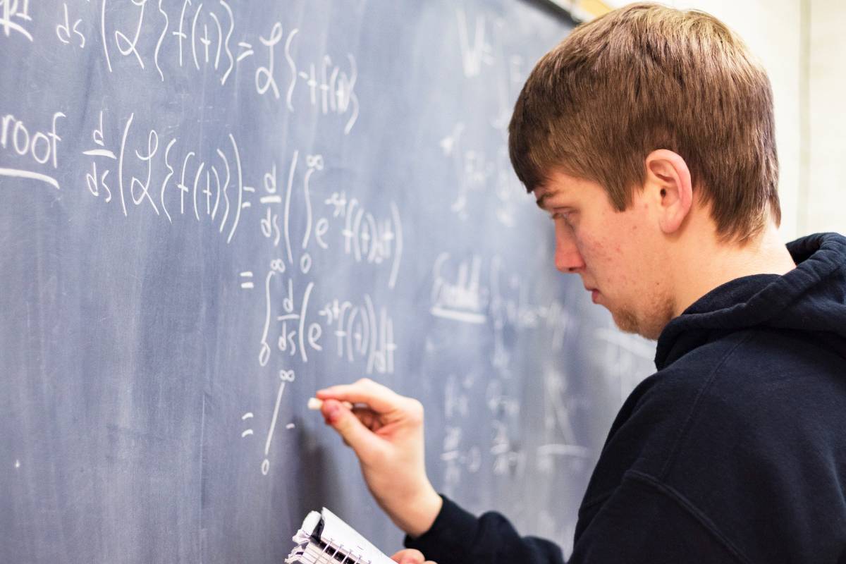 A young man writing advanced calculus equations and proofs on a chalkboard with white chalk.