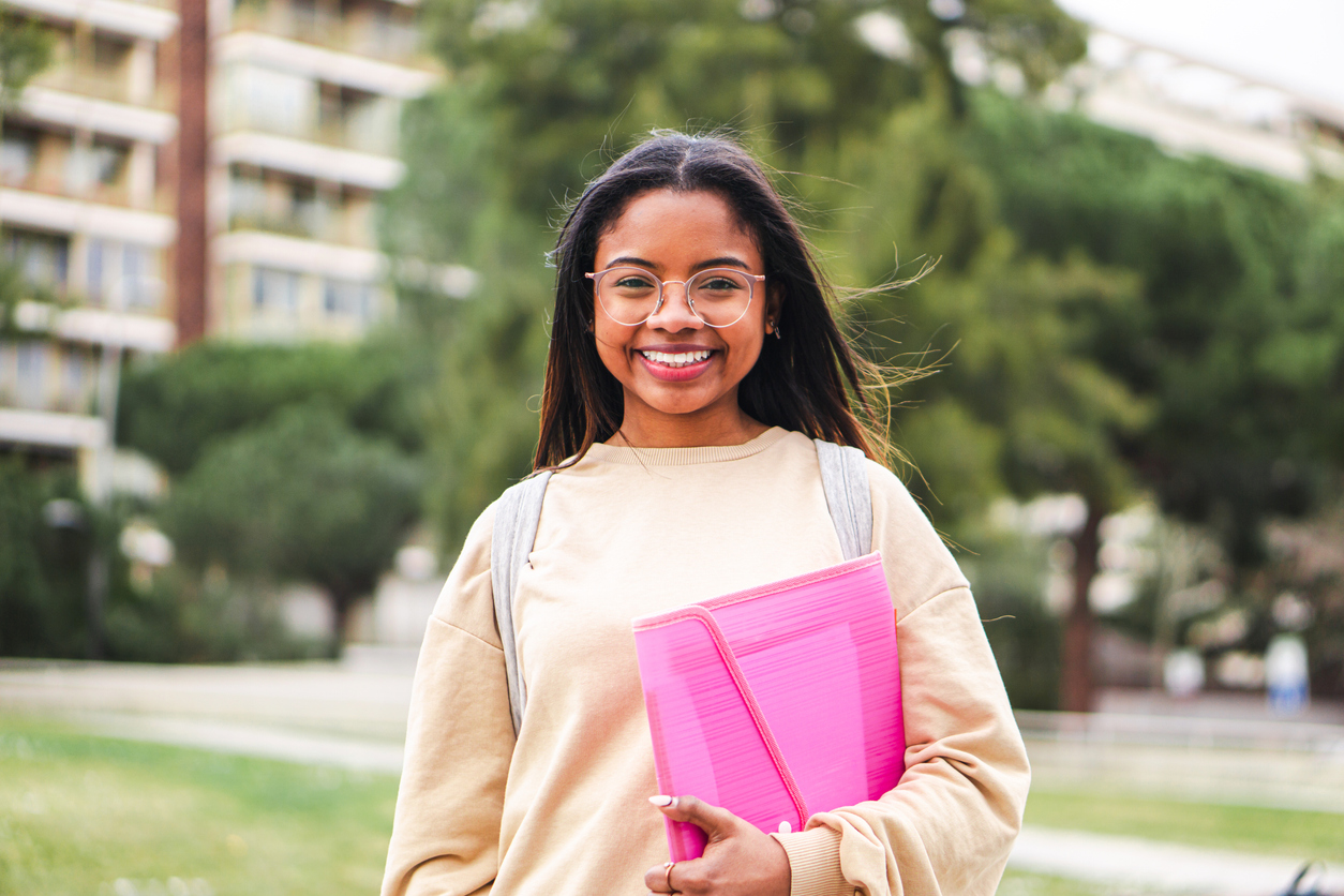 A smiling student wearing glasses and holding a pink folder outdoors with a campus background.