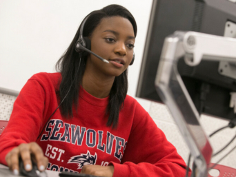 A student wearing a headset sits at a desk and looks at a computer monitor while using a mouse.