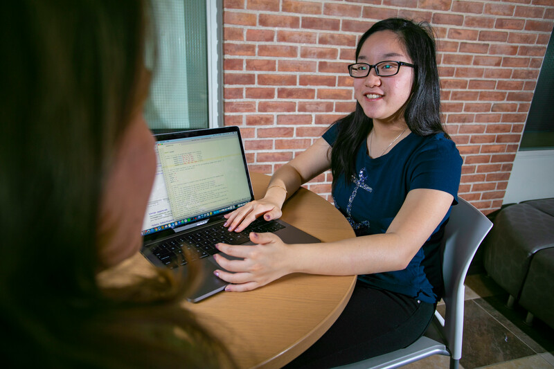 A person sitting at a round table using an open laptop to show code while speaking with another person.