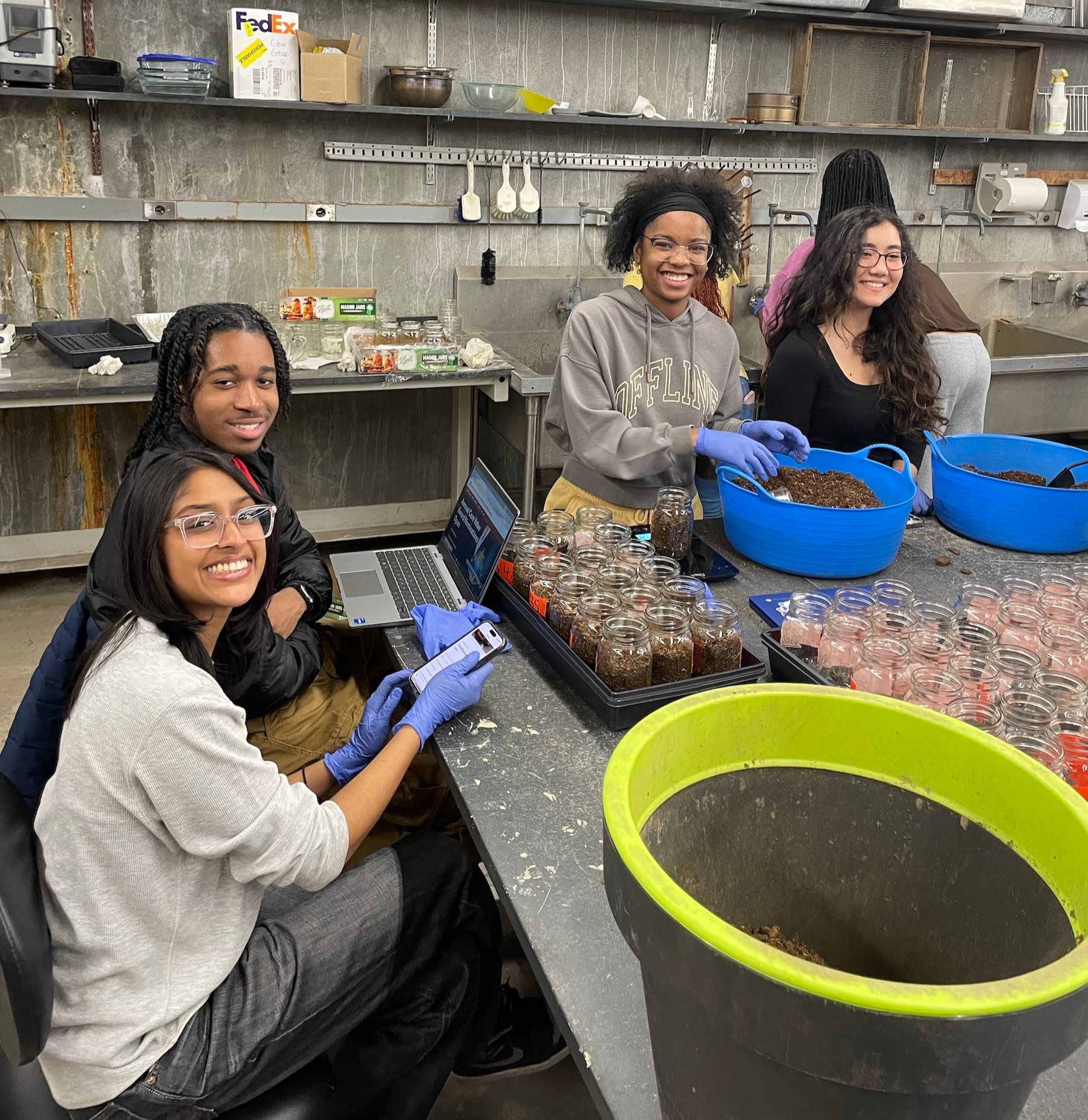 Students working in a laboratory setting with glass jars, soil bins, a laptop, and a smartphone.