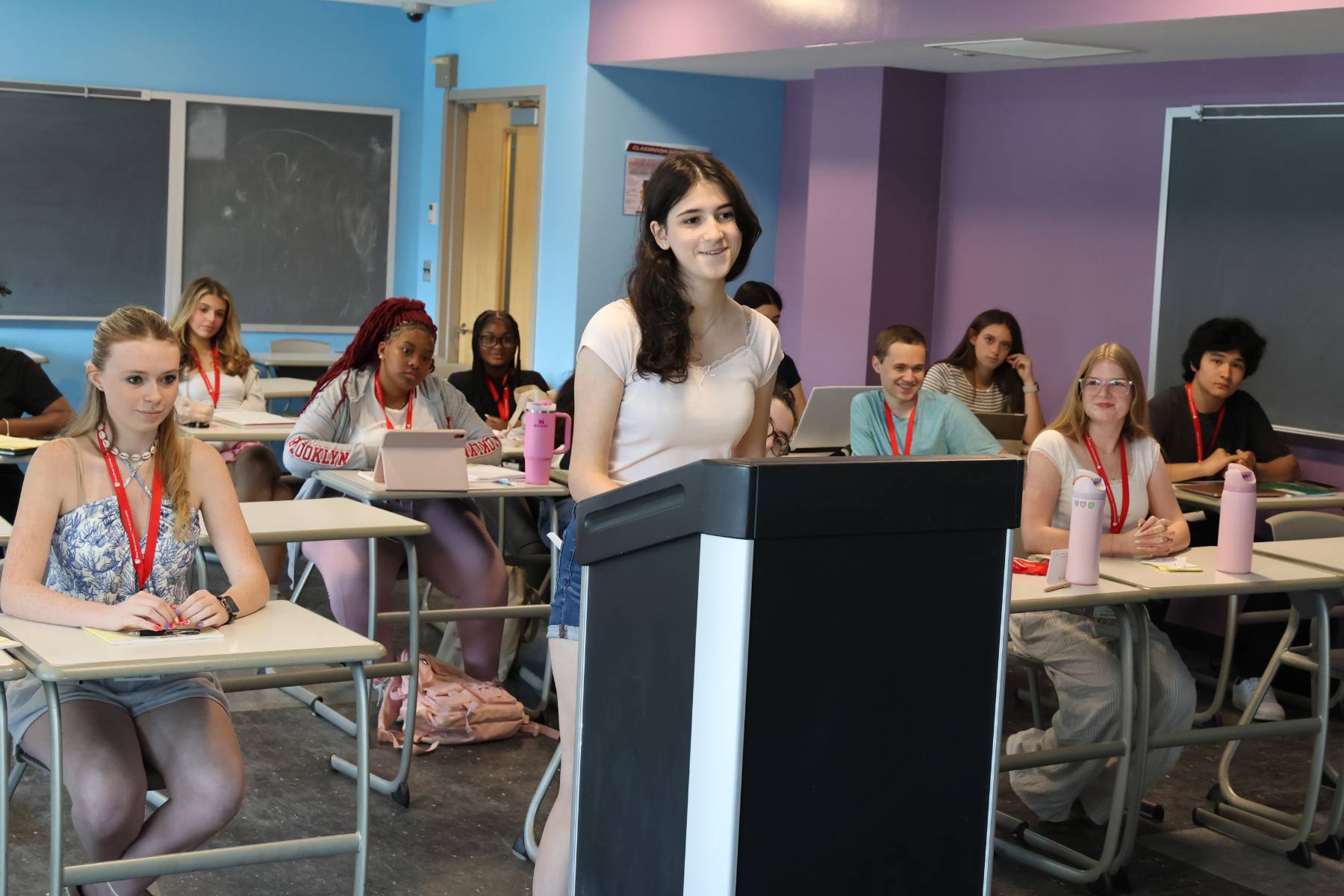 A student stands and speaks at a podium in a classroom while other classmates sit at their desks and listen.