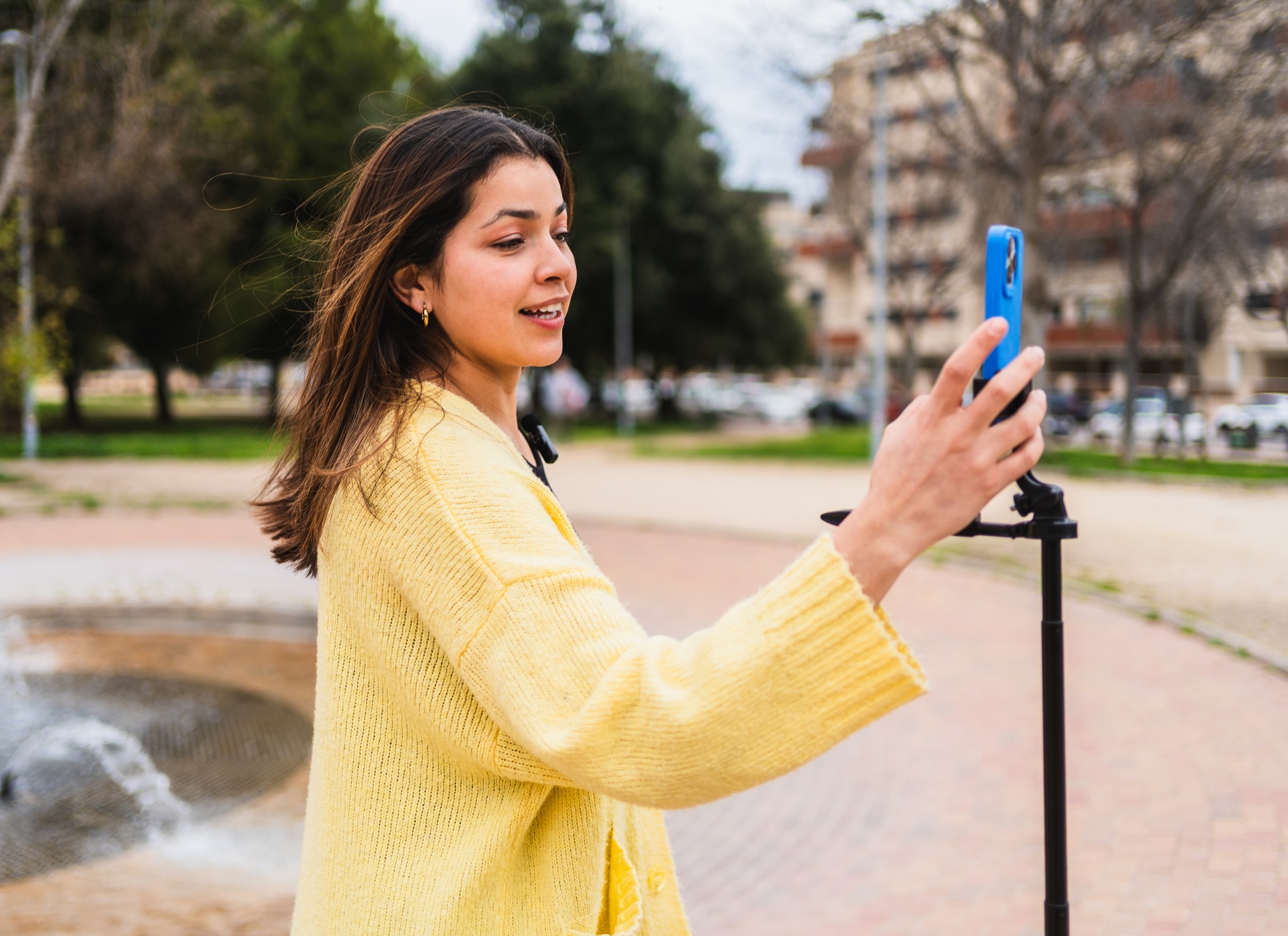 **Alt text:** Person in a yellow sweater recording a video on a smartphone mounted on a tripod in a park.
