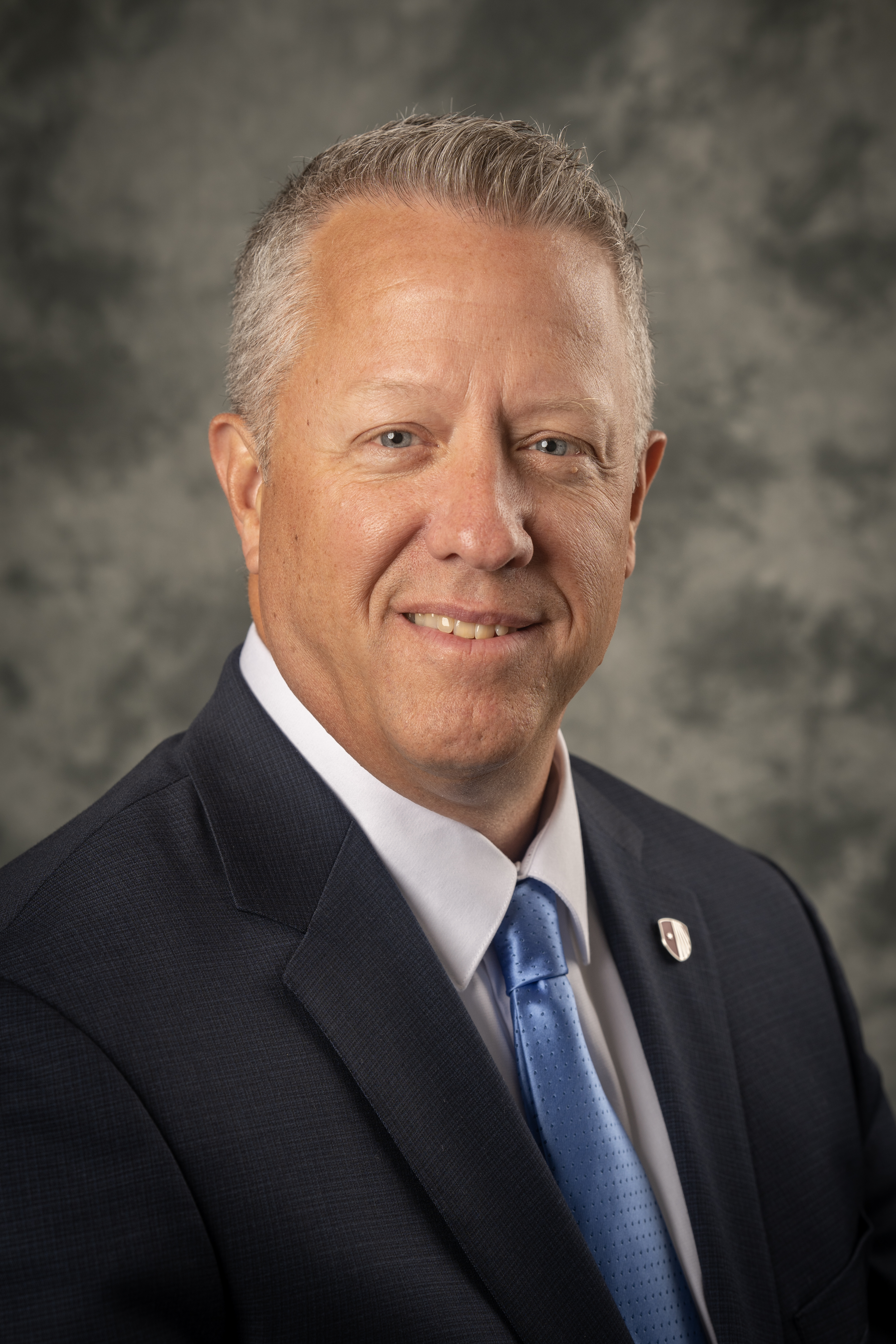 Professional head-and-shoulders portrait of a middle‑aged man with short gray hair, smiling, wearing a dark suit jacket, white shirt, and light blue tie, against a neutral gray studio background.
