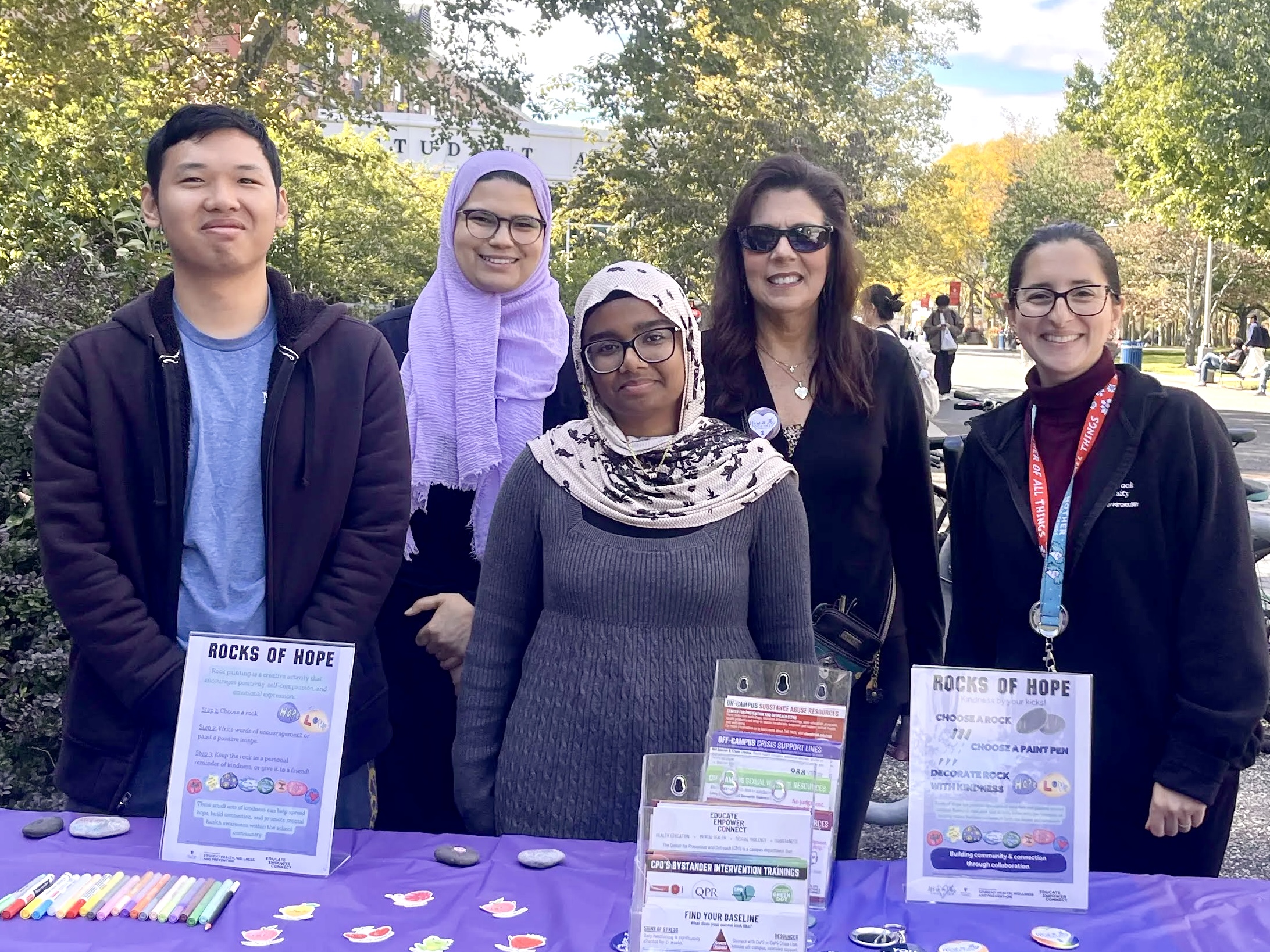 Group photo of five team members behind a table at the 2026 Rocks of Hope event, featuring painted rocks, markers, and informational signs. From left to right: Wesley (PSY 488 Intern), Sarah (Academic Advisor), Nafisa (PSY 488 Intern), Diane (Assistant Director of Undergraduate Programs), and Danielle (Academic Advisor).