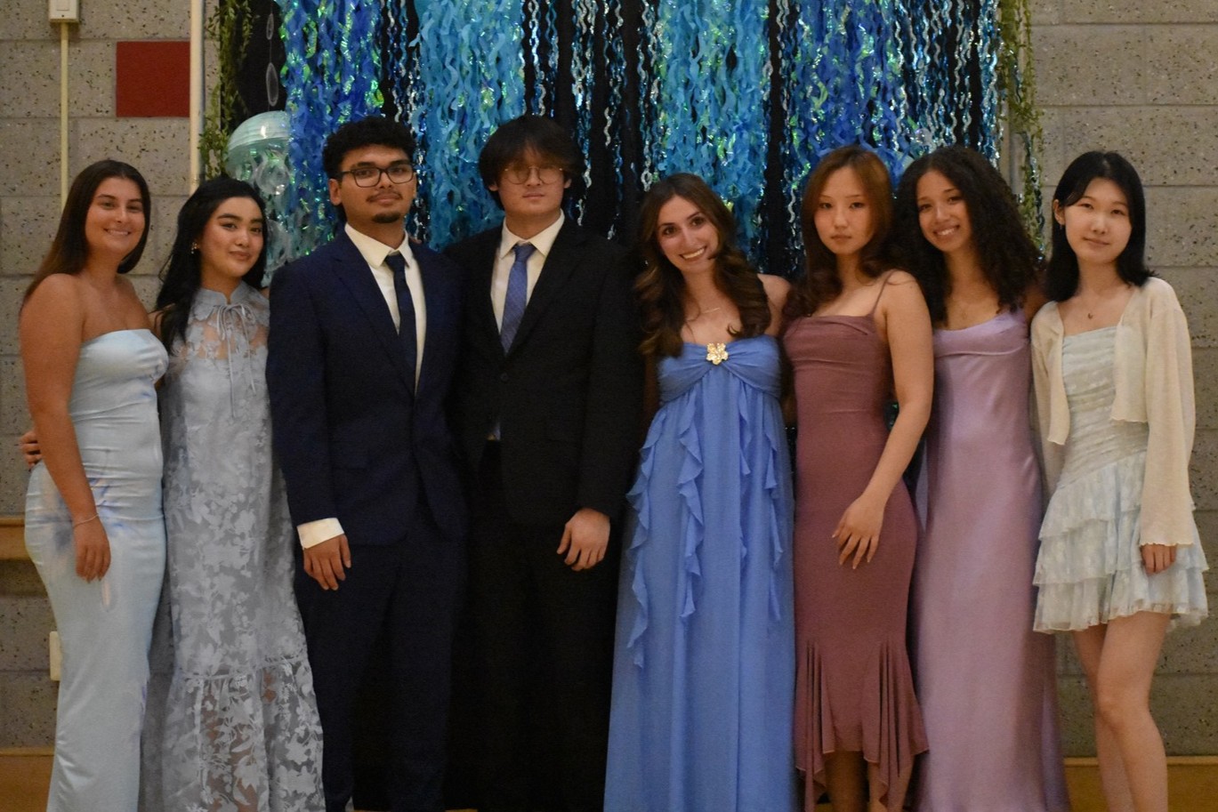 Members of the Stony Brook University Psychology Student Alliance (PSA) Executive Board pose together in formal wear at a gala event in front of a blue and black streamer backdrop.