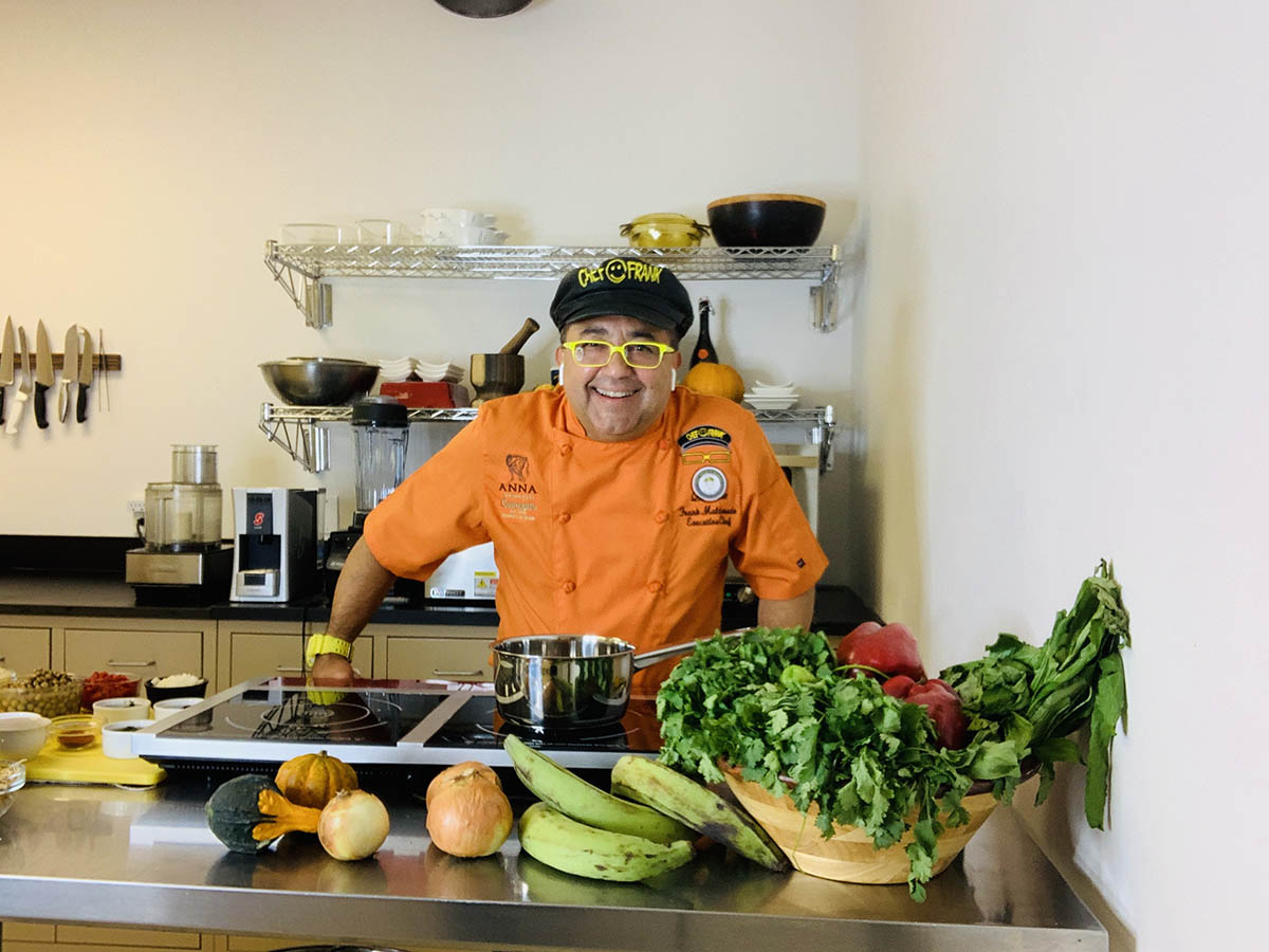 Chef in kitchen wearing orange chef coat standing in front of fruits and vegetables