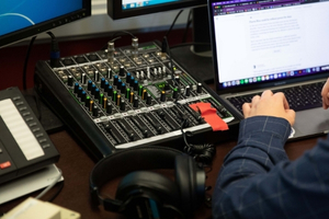 Student operating an audio mixing console and laptop in a broadcast studio.