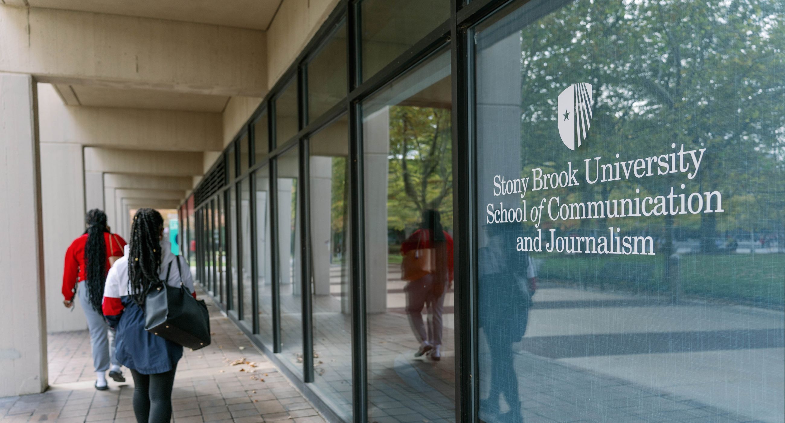 Students walk along a covered campus walkway beside glass windows displaying the Stony Brook University School of Communication and Journalism logo and name.