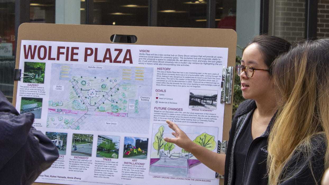 A student stands in front of her Wolfie Plaza poster explaining her project to the visitors