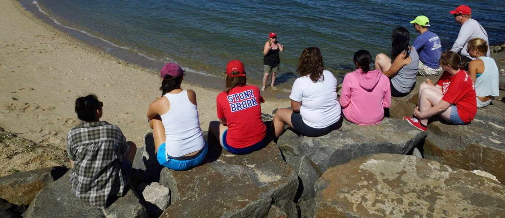 A class listens to their professor while sitting on a jetty at the beach