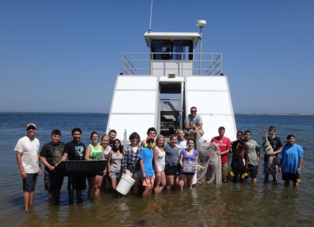 A class of students stand in the water to pose fro a photo in front of the Research Vessel Peconic