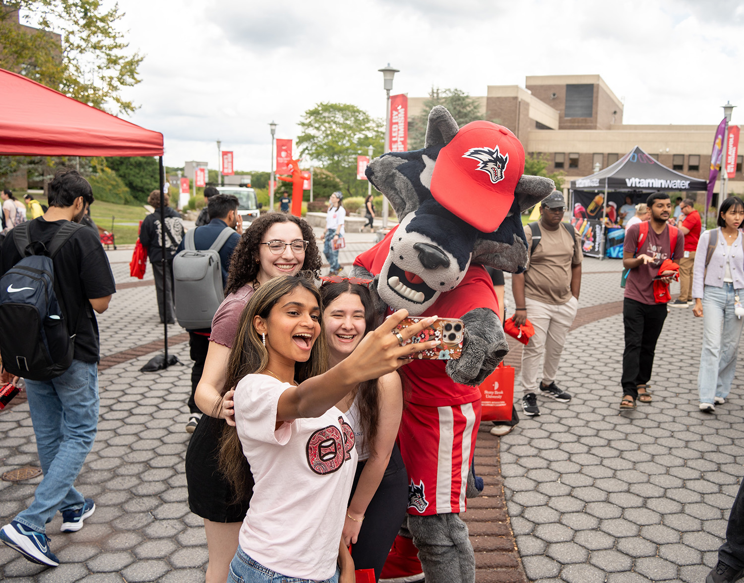 Students talking a selfie with a campus mascot at an outdoor event.