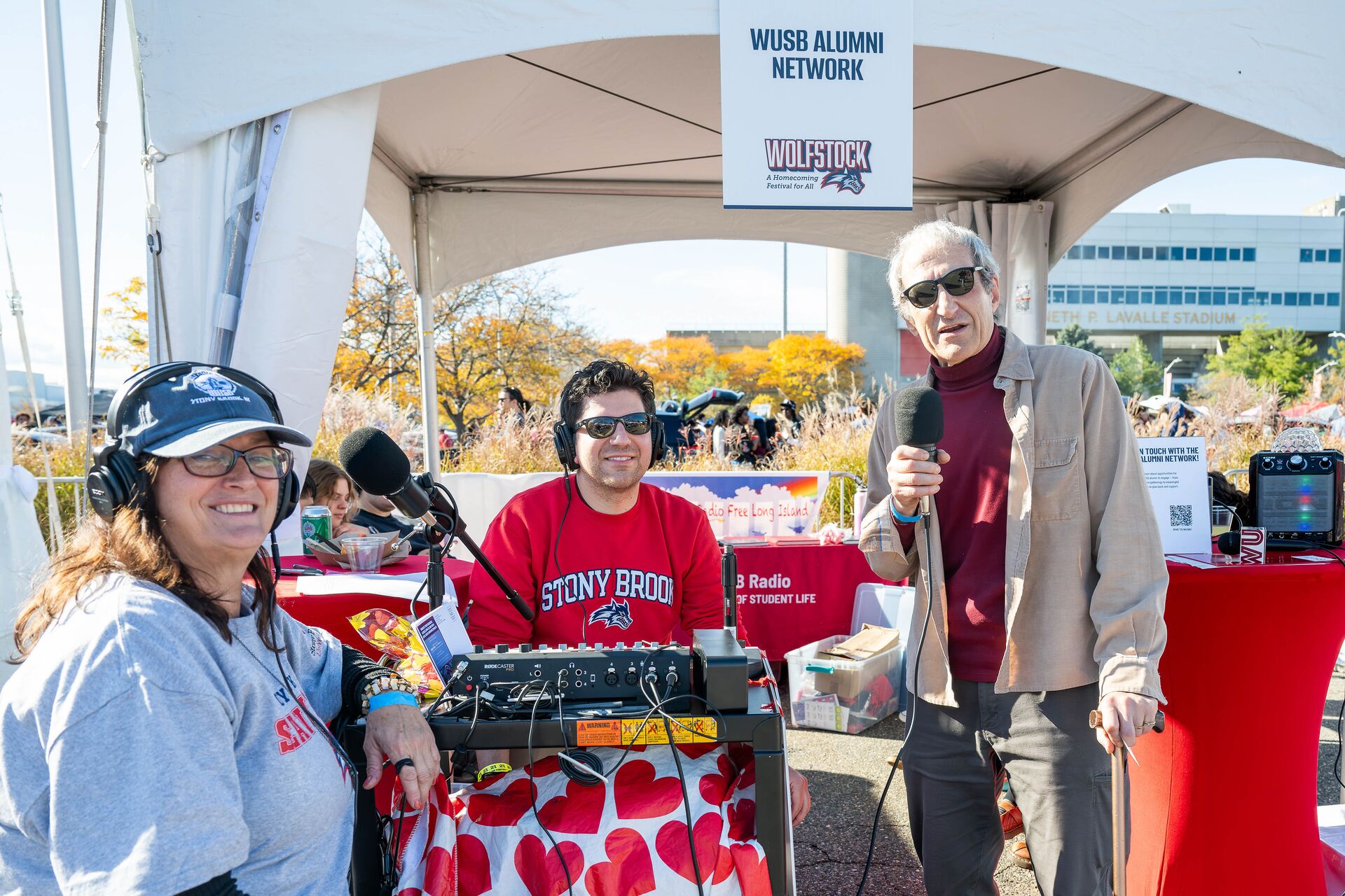 Two people sitting at an outdoor table with a recording setup. An older man stands next to them with a microphone.