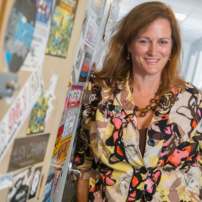 A smiling woman leaning against lockers covered in radio station stickers.
