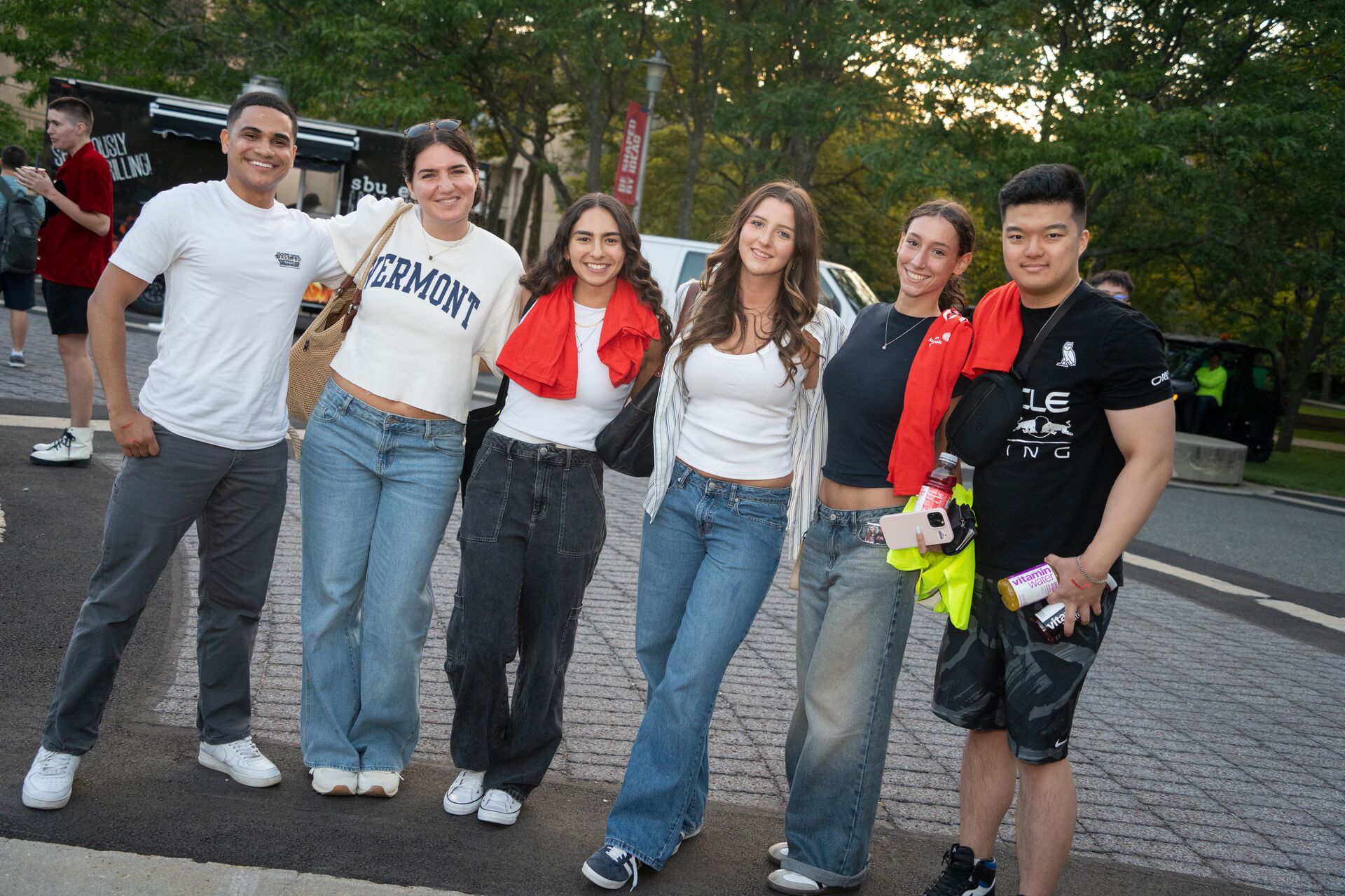 Six people stand arm in arm and smile for a photo on a campus plaza during an evening event, with red event T-shirts draped over their shoulders and trees and other attendees in the background.