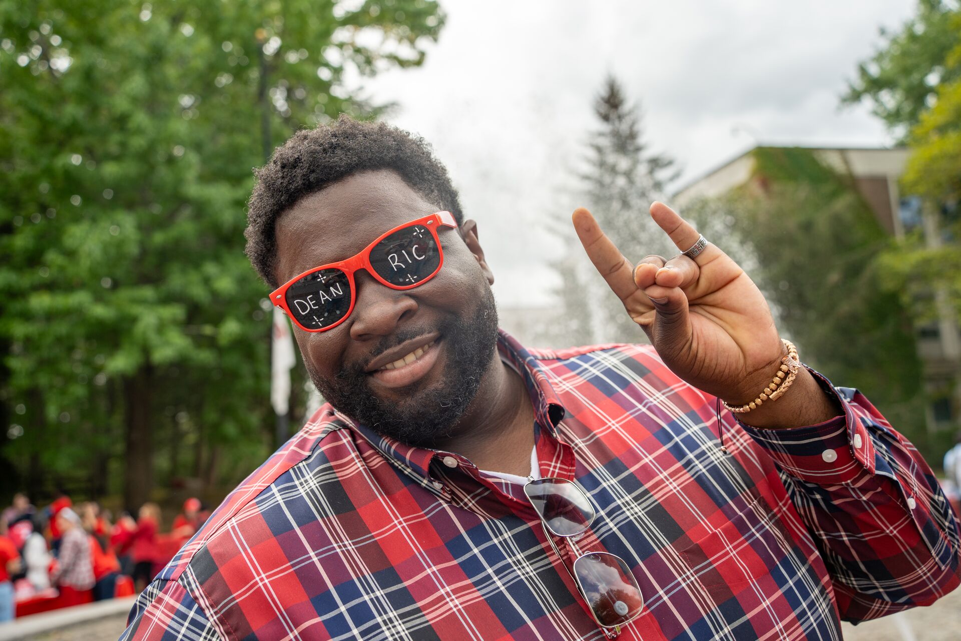 Smiling portrait of a man wearing red novelty sunglasses that read "DEAN RIC" while making a university hand sign outdoors.