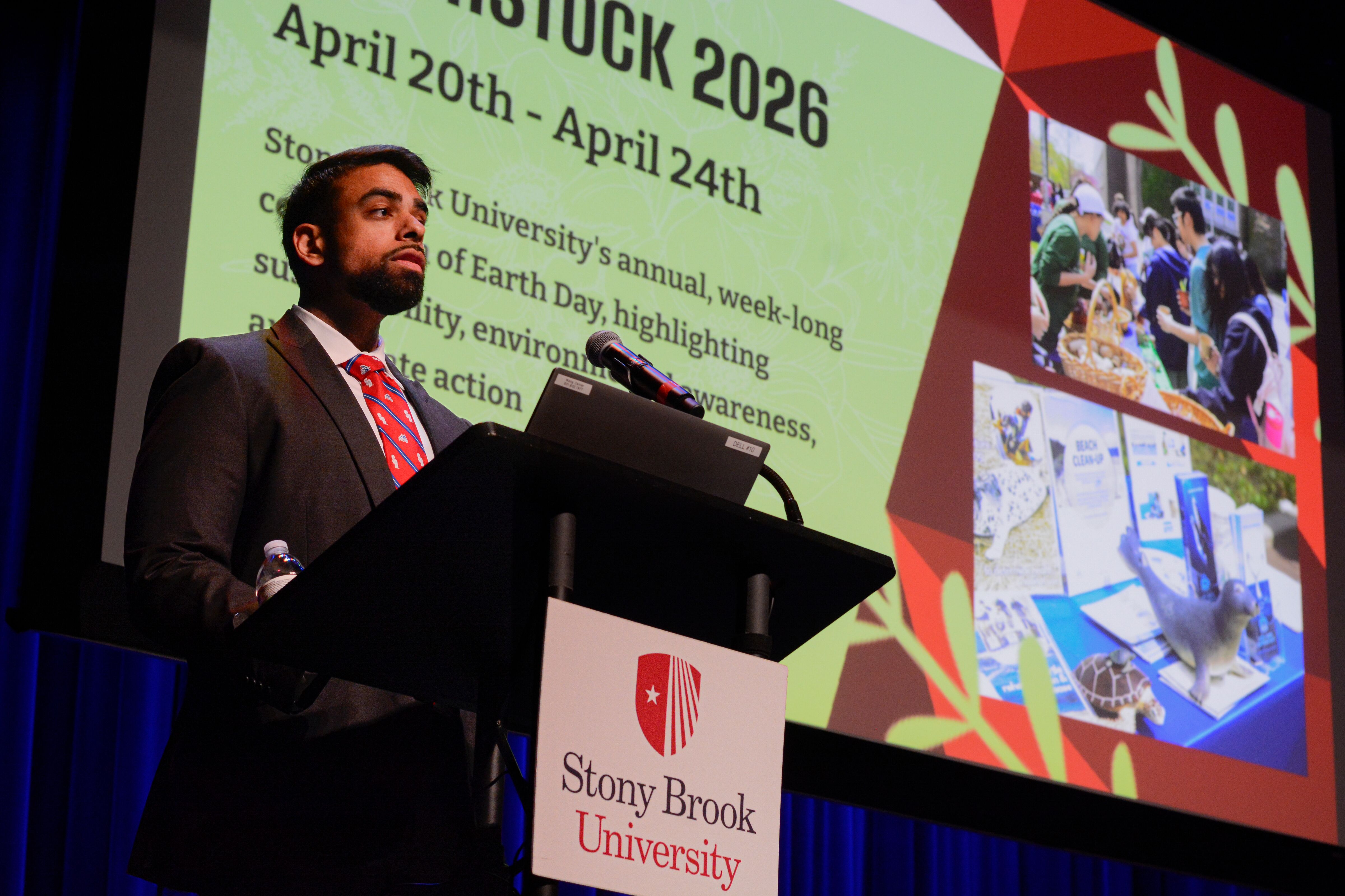 A presenter speaks at a podium in front of a projector screen displaying "Earthstock 2026" text and campus event photos.