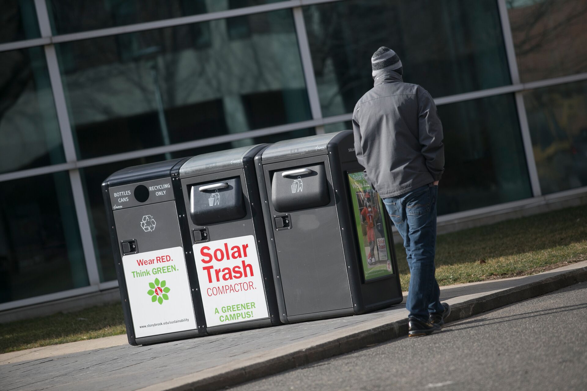 A person walks past a row of outdoor solar-powered trash and recycling compactors on a university campus.