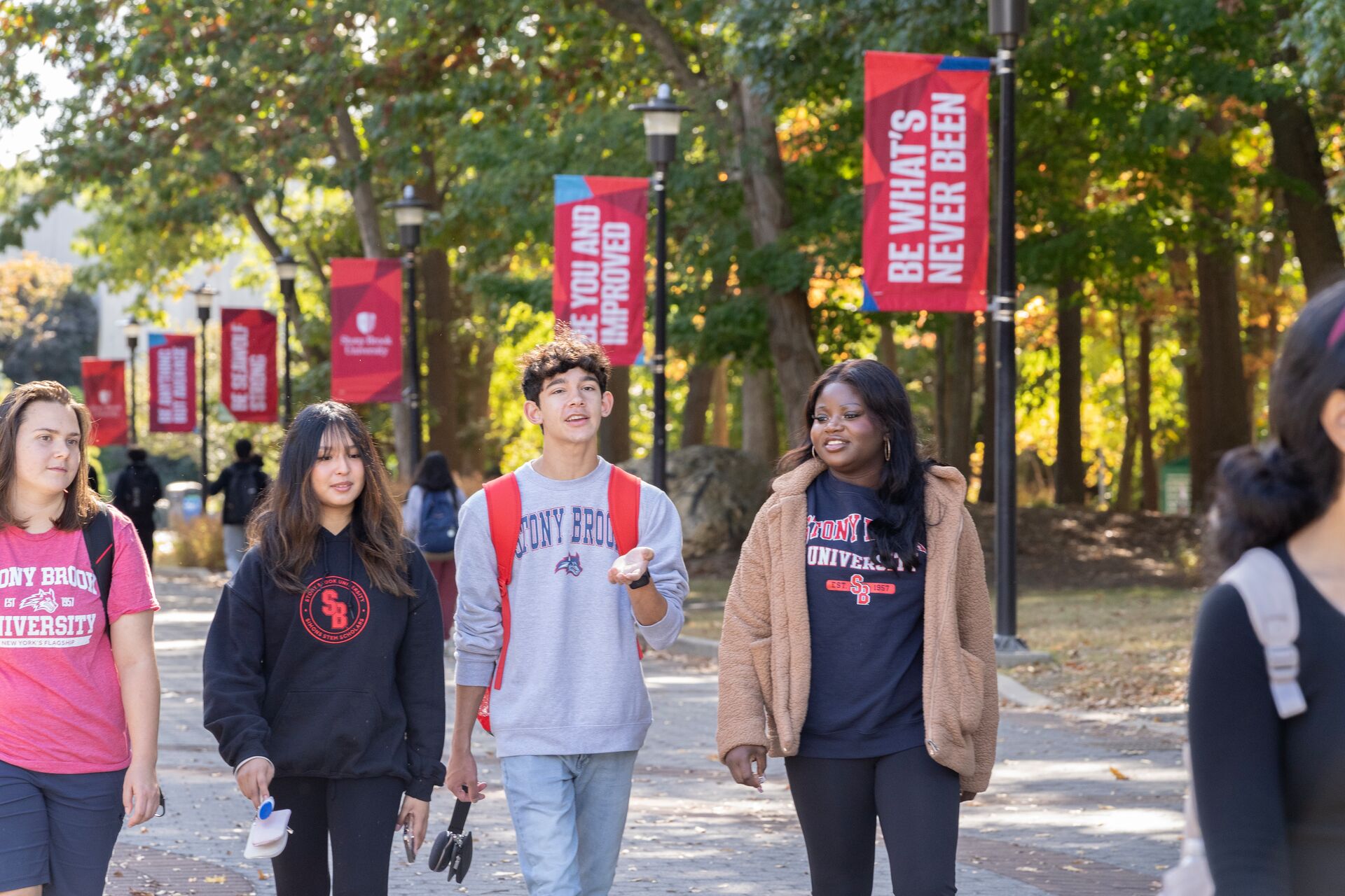 A red campus lamppost banner reading "BE UNDENIABLE" in white vertical text, framed by green tree leaves.