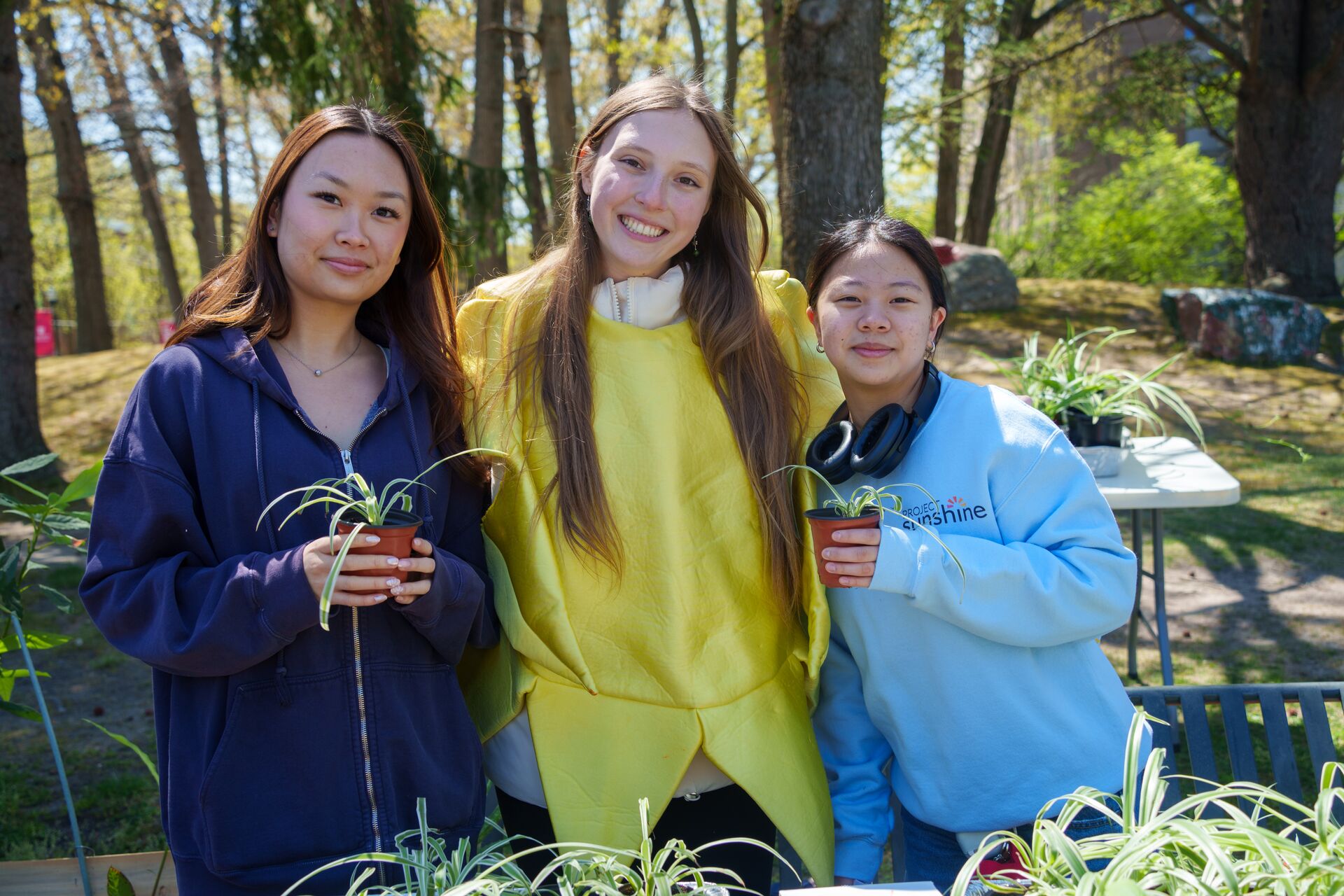 Three female students stand shoulder-to-shoulder in a wooded campus area. The student in the center wears a bright yellow, plush banana costume. The two students flanking her hold up tiny plastic pots containing small spider plant starts.