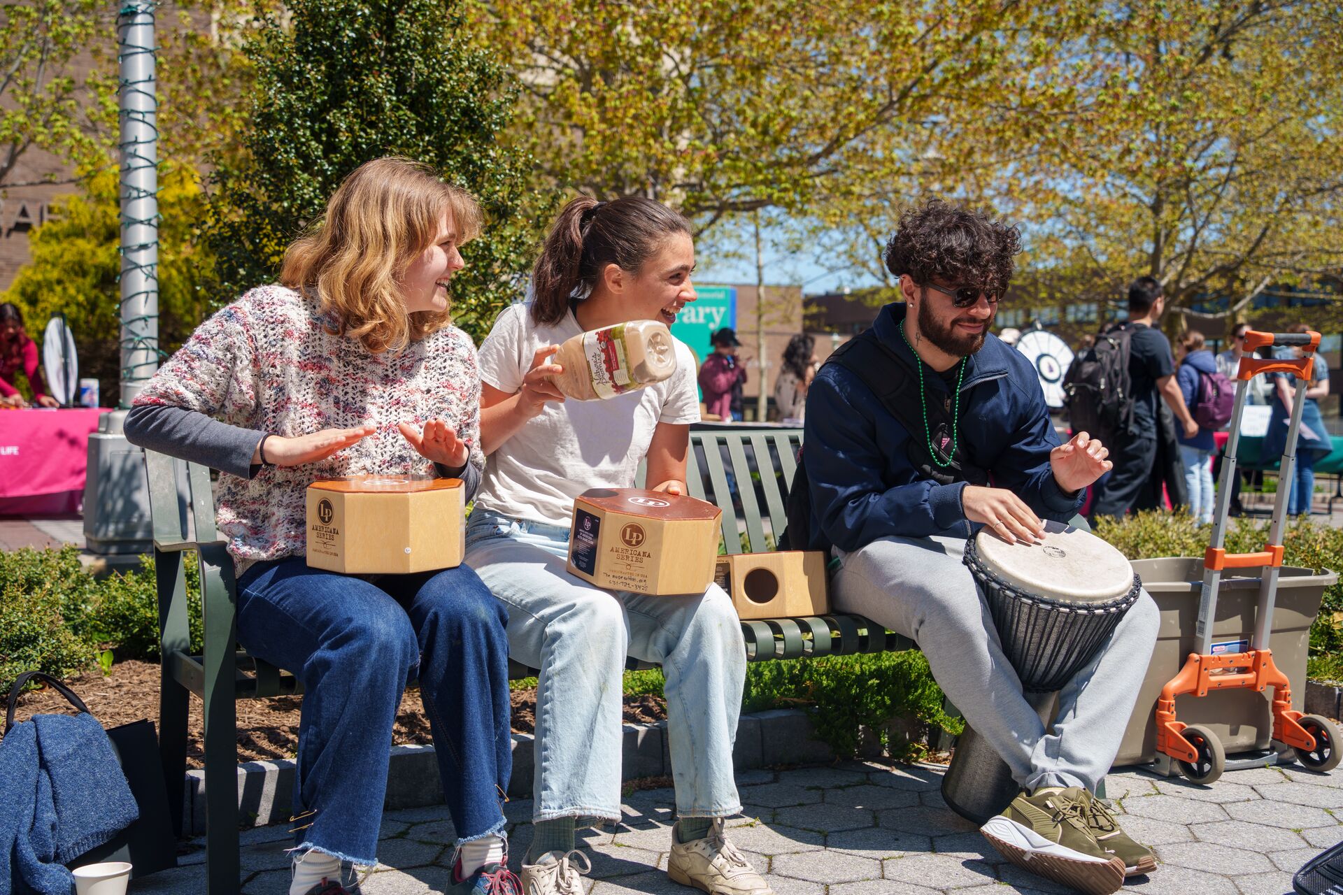Three students sit on a campus bench playing hand drums and a shaker during an outdoor festival.