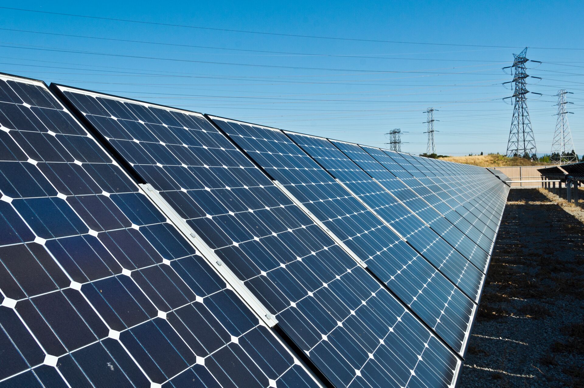 A long, angled row of blue photovoltaic solar panels under a clear blue sky, with power lines and transmission towers in the background.
