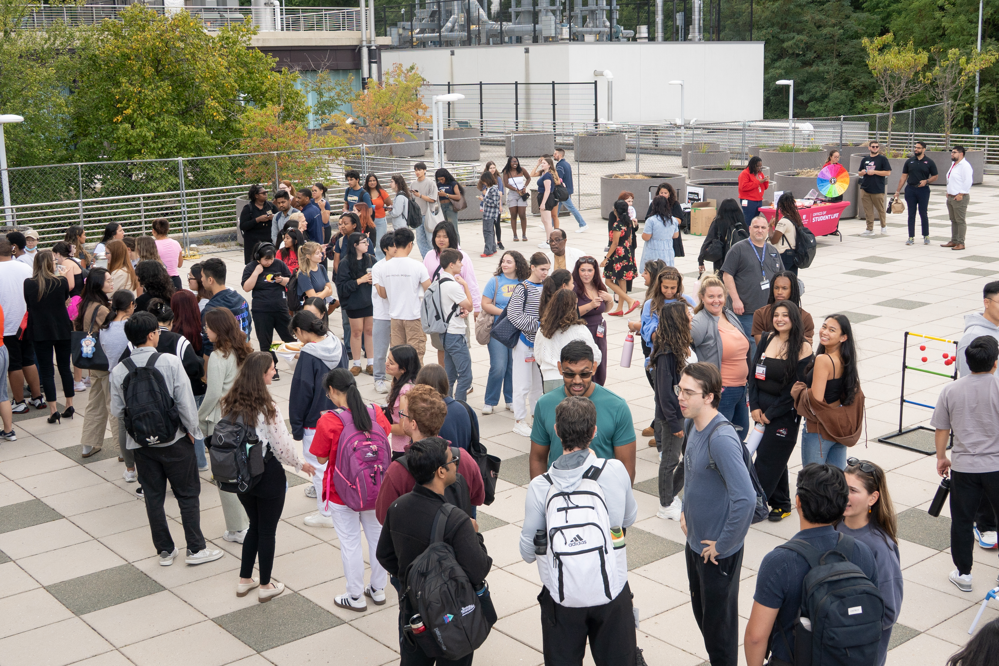 A cheerful group of students, including a few wearing "Student Nurse" ID badges, stand together outside on a tiled campus patio. They are talking and laughing genuinely, with a ladder ring toss game visible on the right.