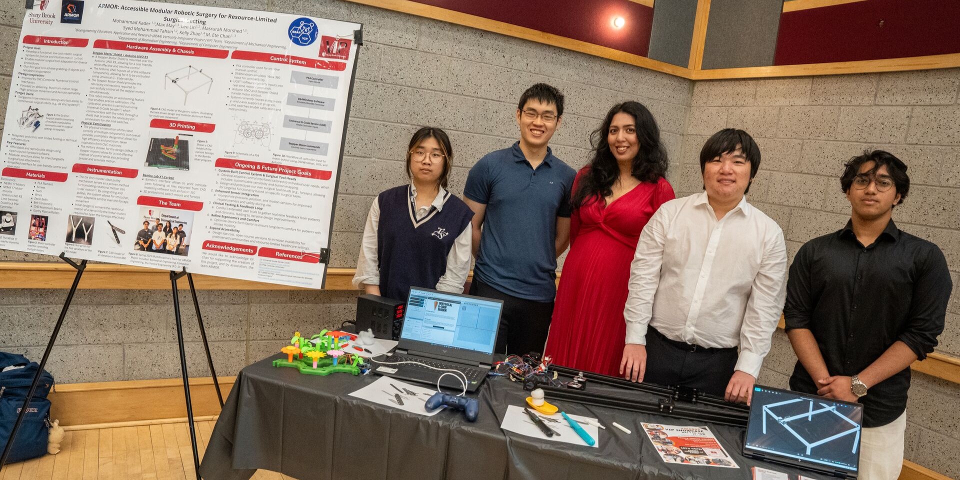 Five students standing behind a desk with computers and gadgets. There is a poster to the left of the desk displaying research.
