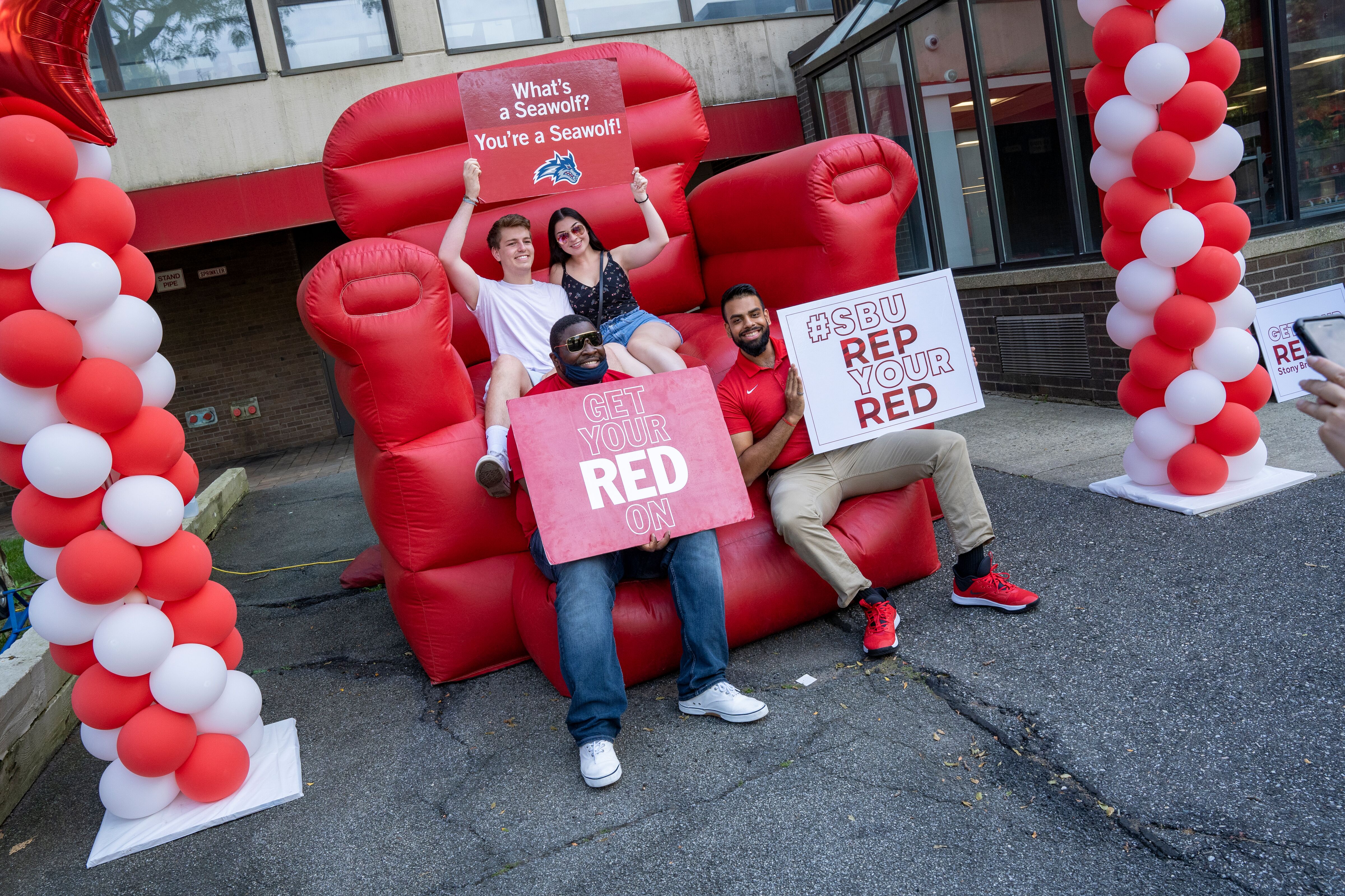 Four people sit on an inflatable red chair holding signs reading "SBU rep your red"